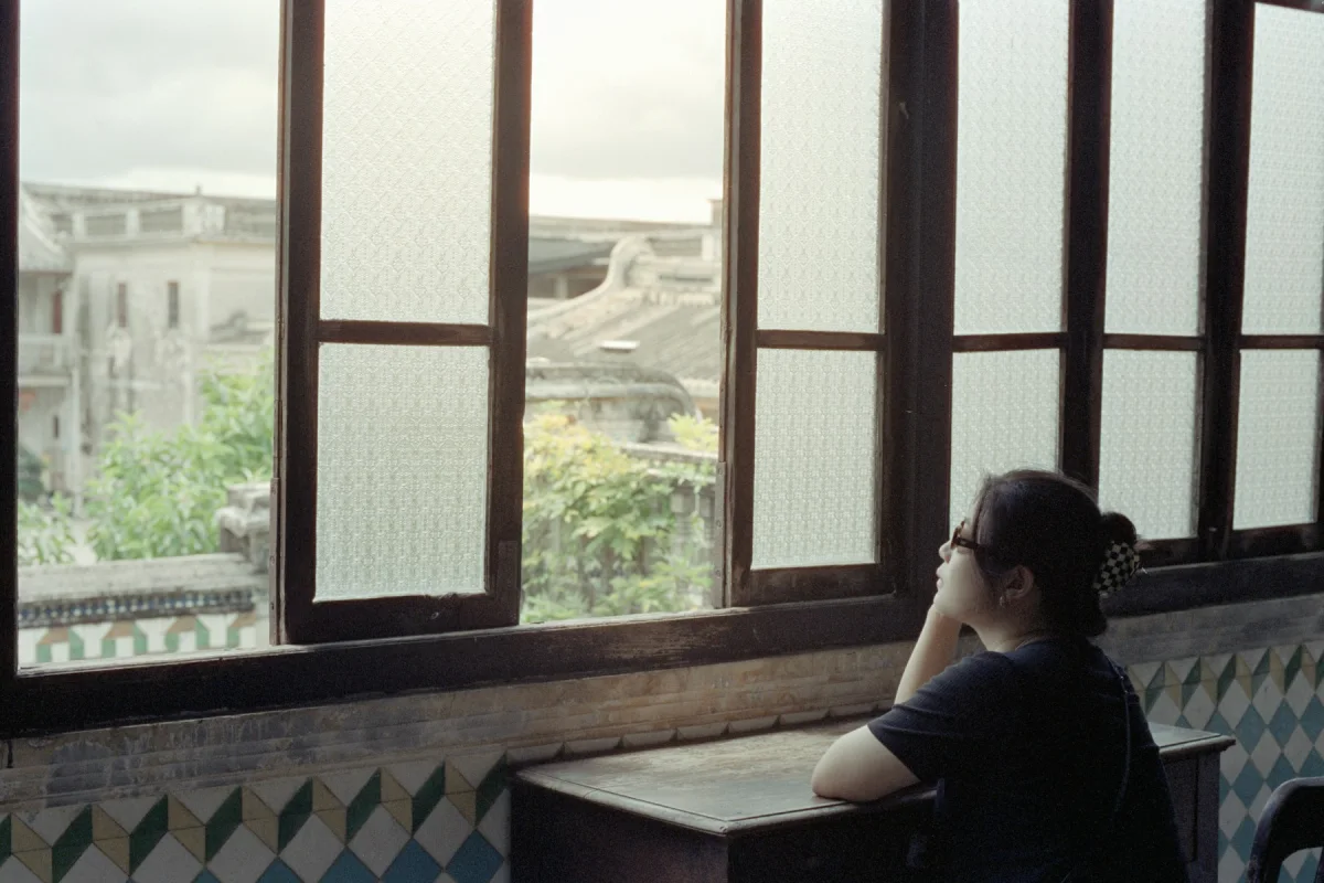 A woman sitting quietly beside an old window and looking out across neighbouring rooftops